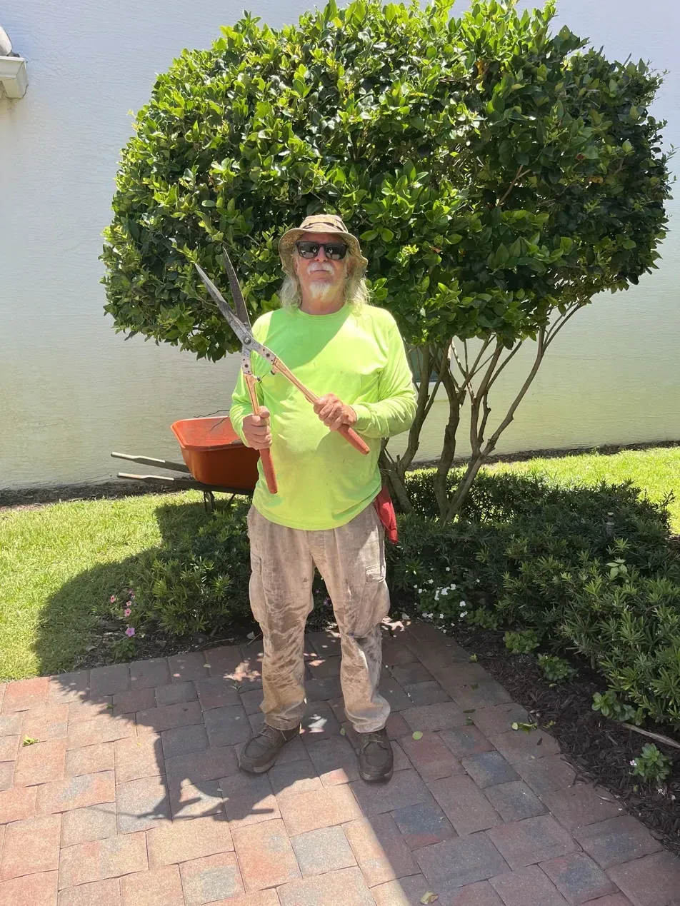 Tim, owner of Tim's Lawn Service, holding hedge shears in front of a freshly trimmed tree and mulched beds